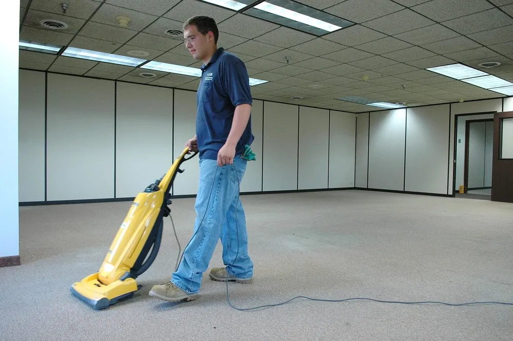 A cleaner running an upright vacuum across the carpet of an empty office floor